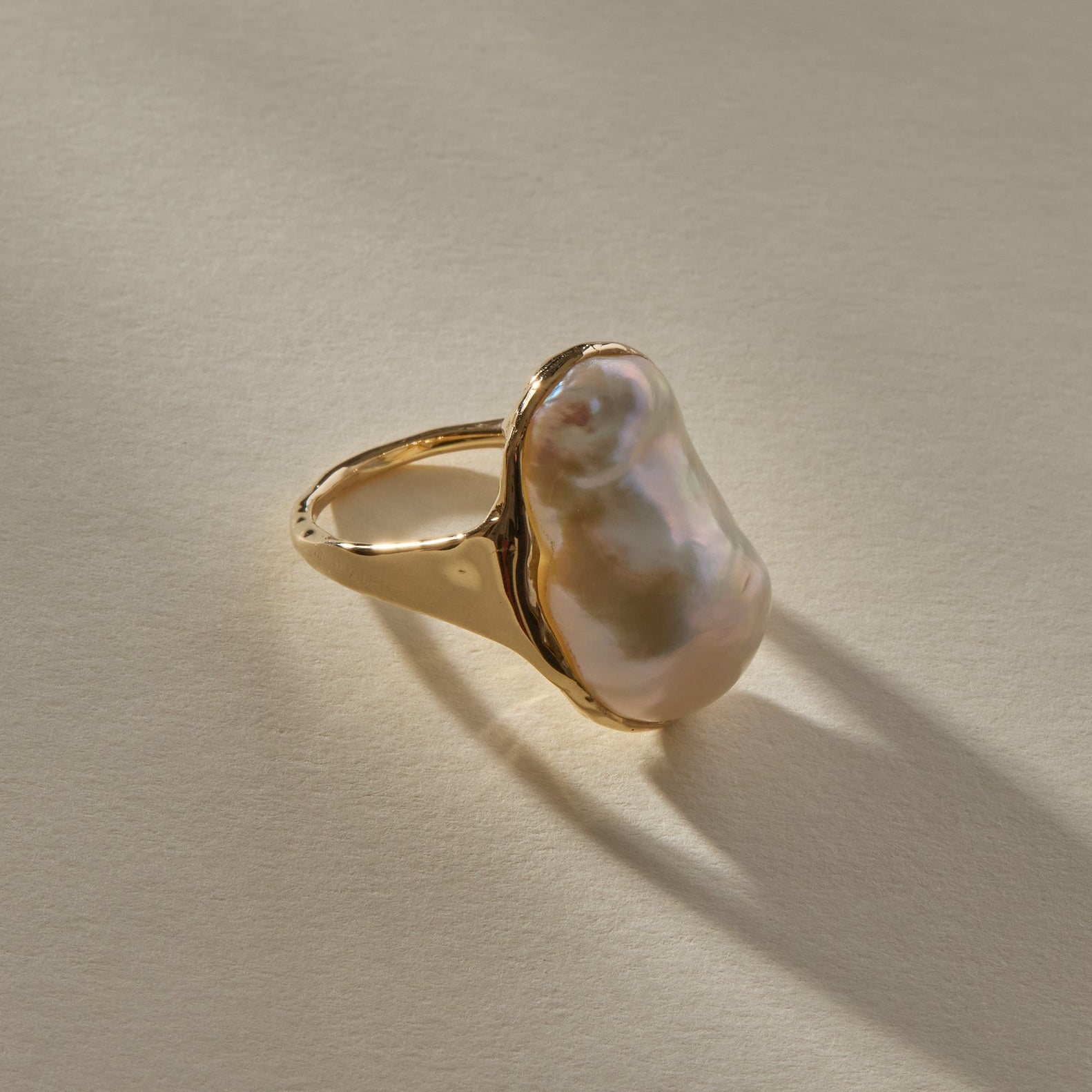 A gold signet-style ring featuring a large, irregular baroque pearl, photographed on a textured beige surface with a soft shadow.