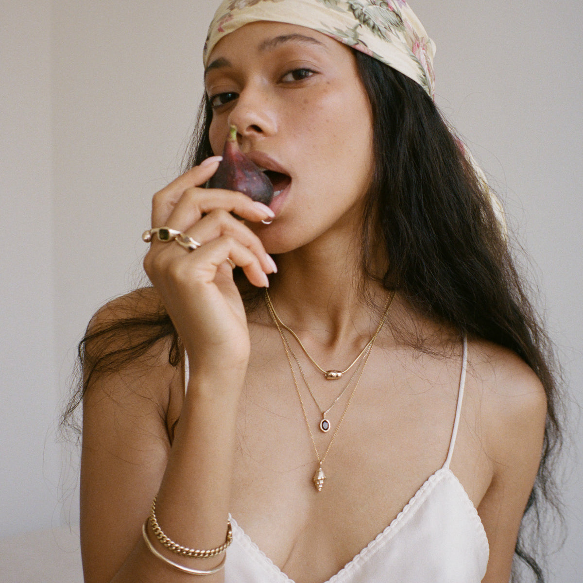 Woman wearing a floral headscarf and gold jewelry, holding a small object near her mouth.