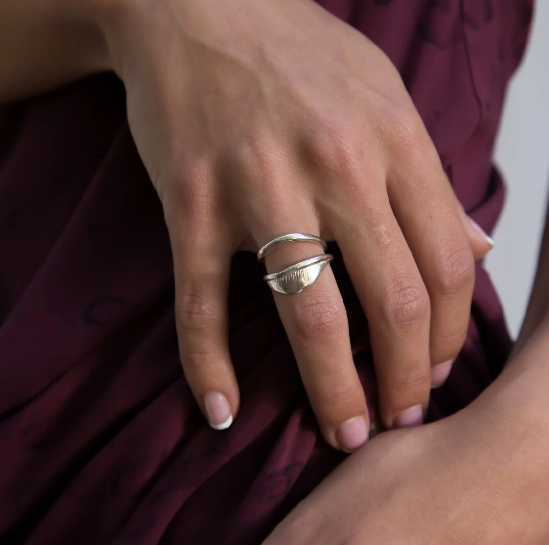 Hand resting on burgundy fabric wearing the Devotions Wave Ring stacked with another ring.