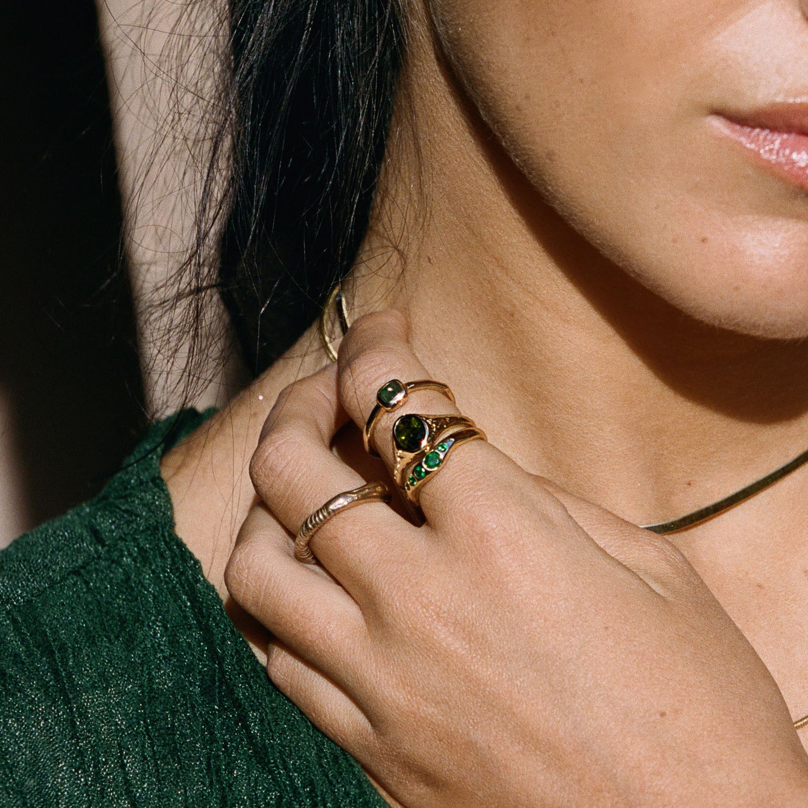 Model in a green top with her hand at her neck, wearing the Eros Five-Stone Emerald Band with stacked rings.