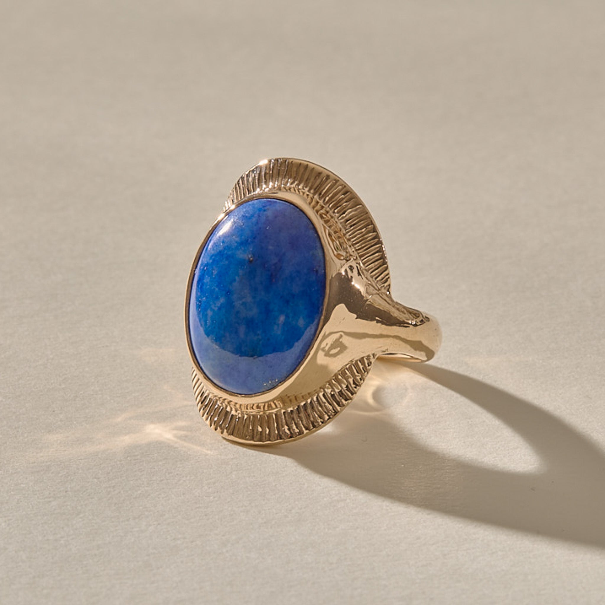 Inanna Lapis Shield Ring in solid recycled gold, an oval Chilean lapis lazuli cabochon framed by a fine carved sunburst setting, in warm sunlight.