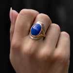 A hand against a black background wearing the Inanna Lapis Shield Ring, the blue lapis cabochon facing forward.
