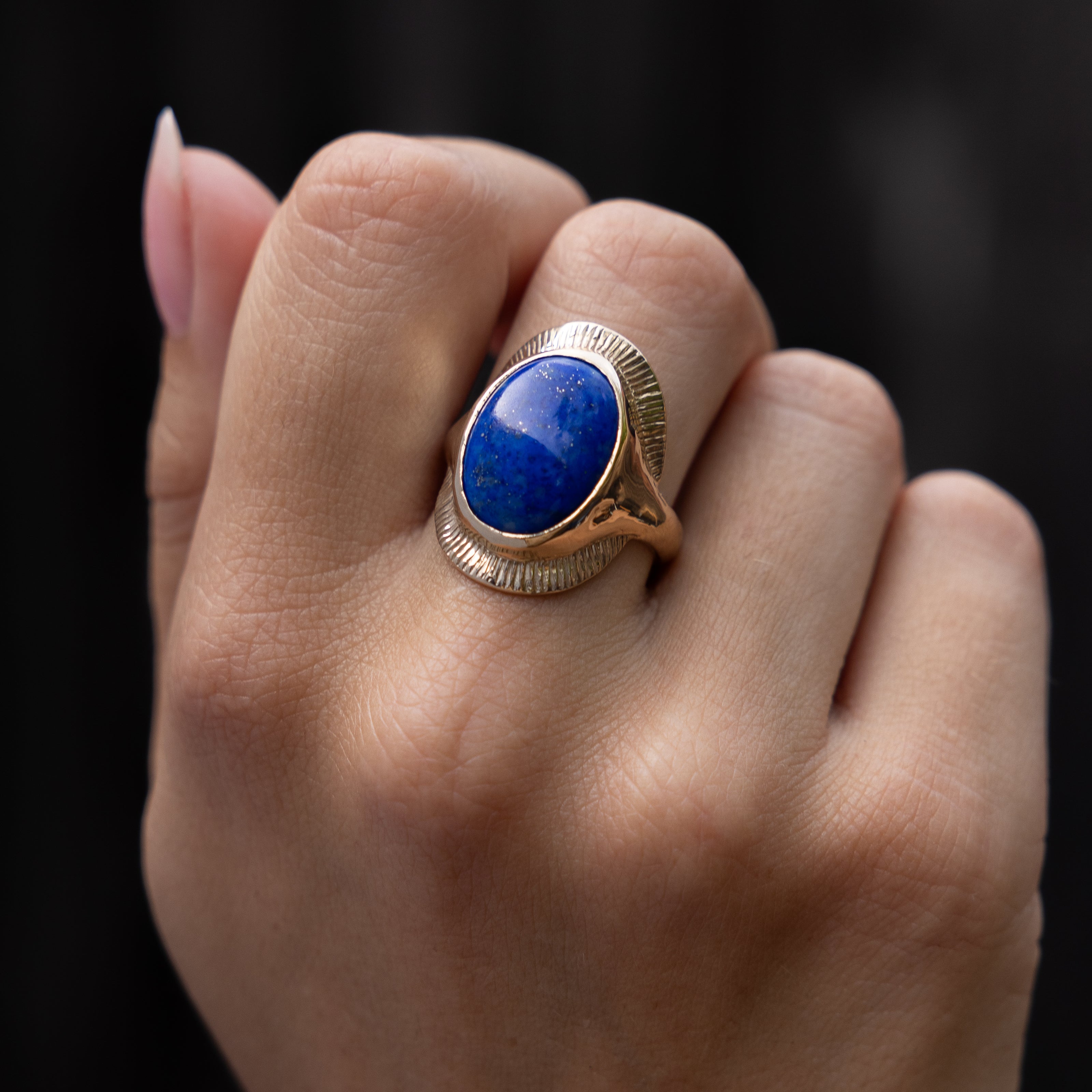 A hand against a black background wearing the Inanna Lapis Shield Ring, the blue lapis cabochon facing forward.