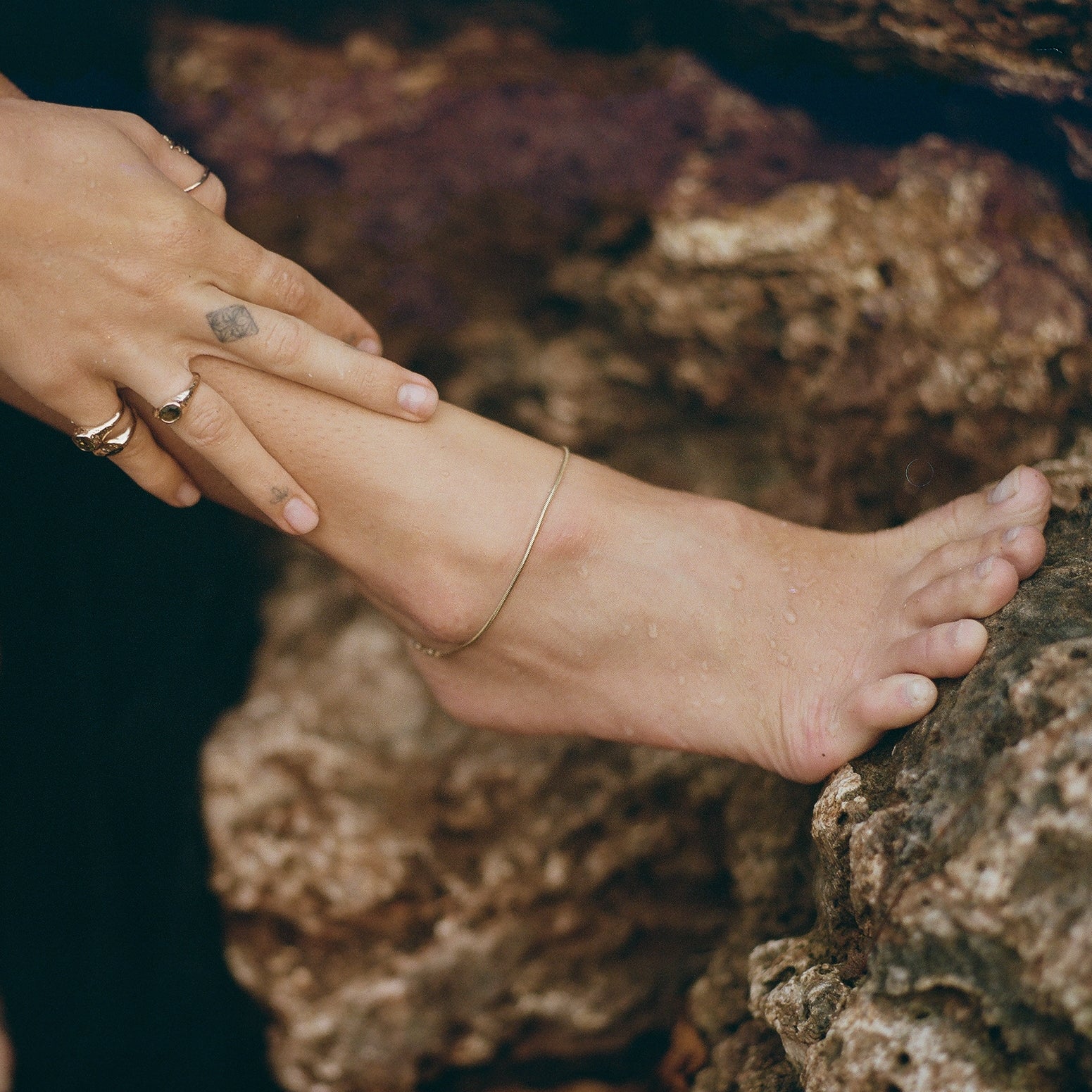 Hand with gold rings resting on a foot wearing the gold Jera Snake Chain Anklet on dark rocks.