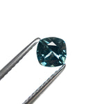 The cushion cut Montana sapphire held in tweezers, showing its cool blue-teal color and sparkle.