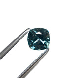 The cushion cut Montana sapphire held in tweezers, showing its cool blue-teal color and sparkle.
