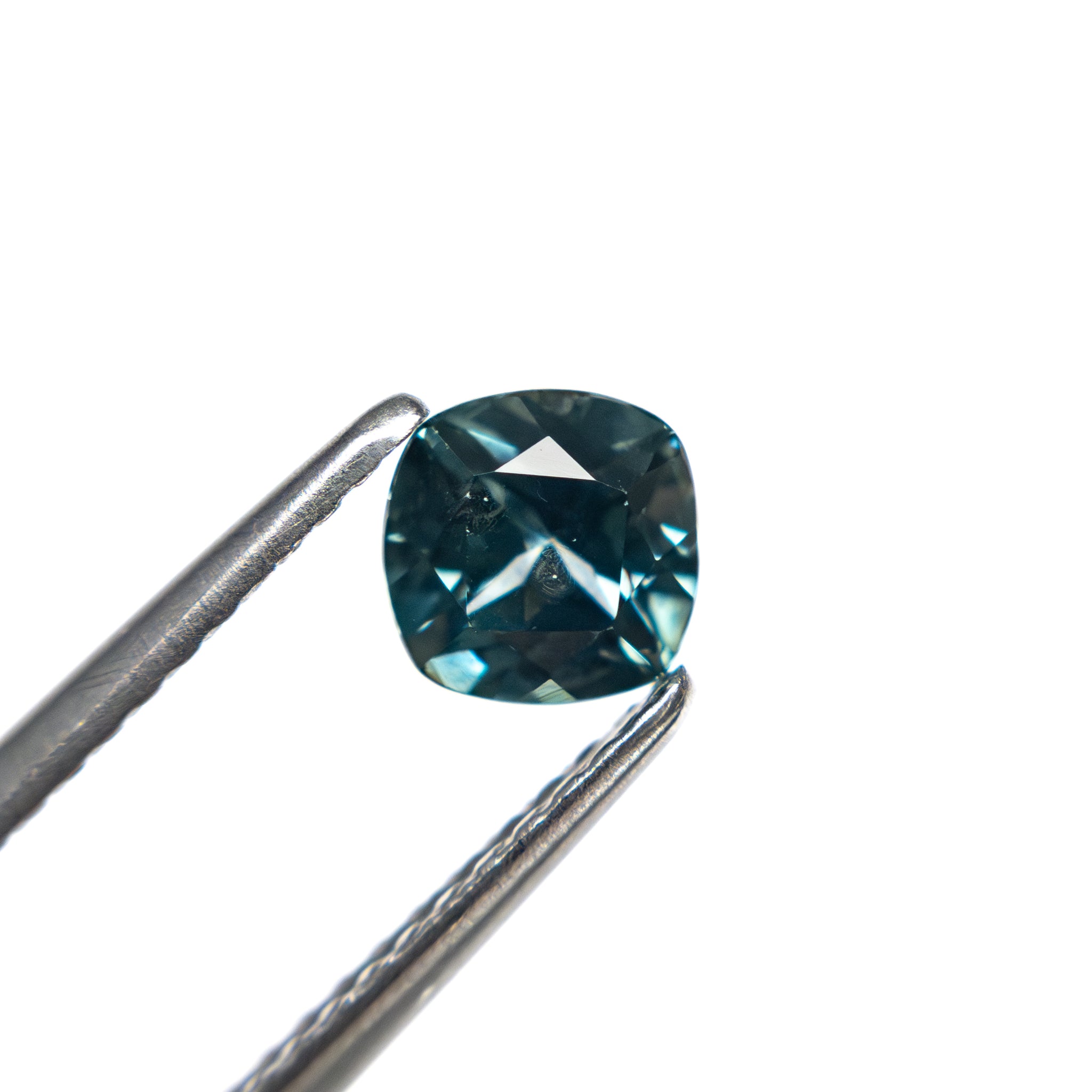 The cushion cut Montana sapphire held in tweezers, showing its cool blue-teal color and sparkle.