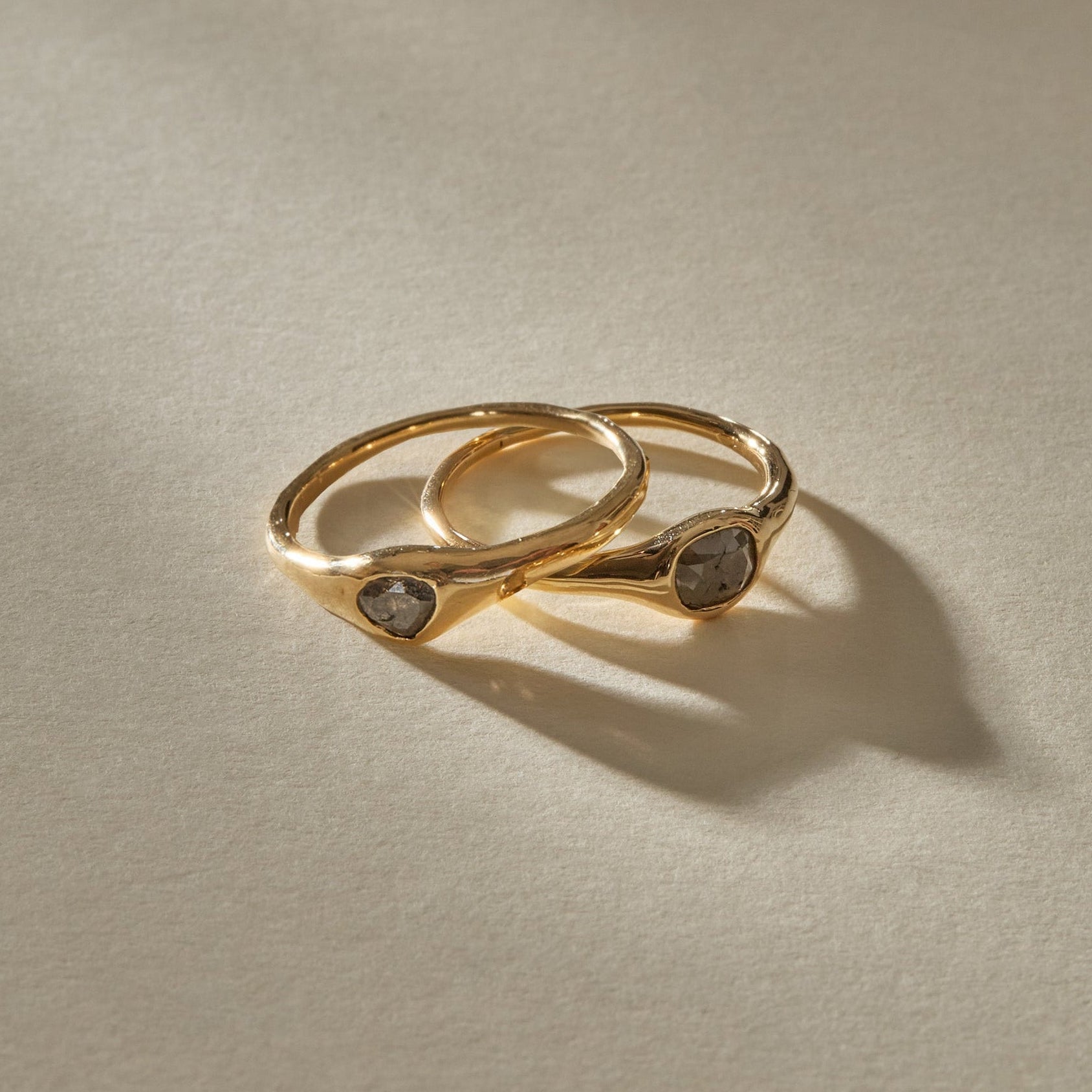 The Selkie Stack II, two one of a kind natural grey diamond rings in organic 14k recycled gold settings, resting together.