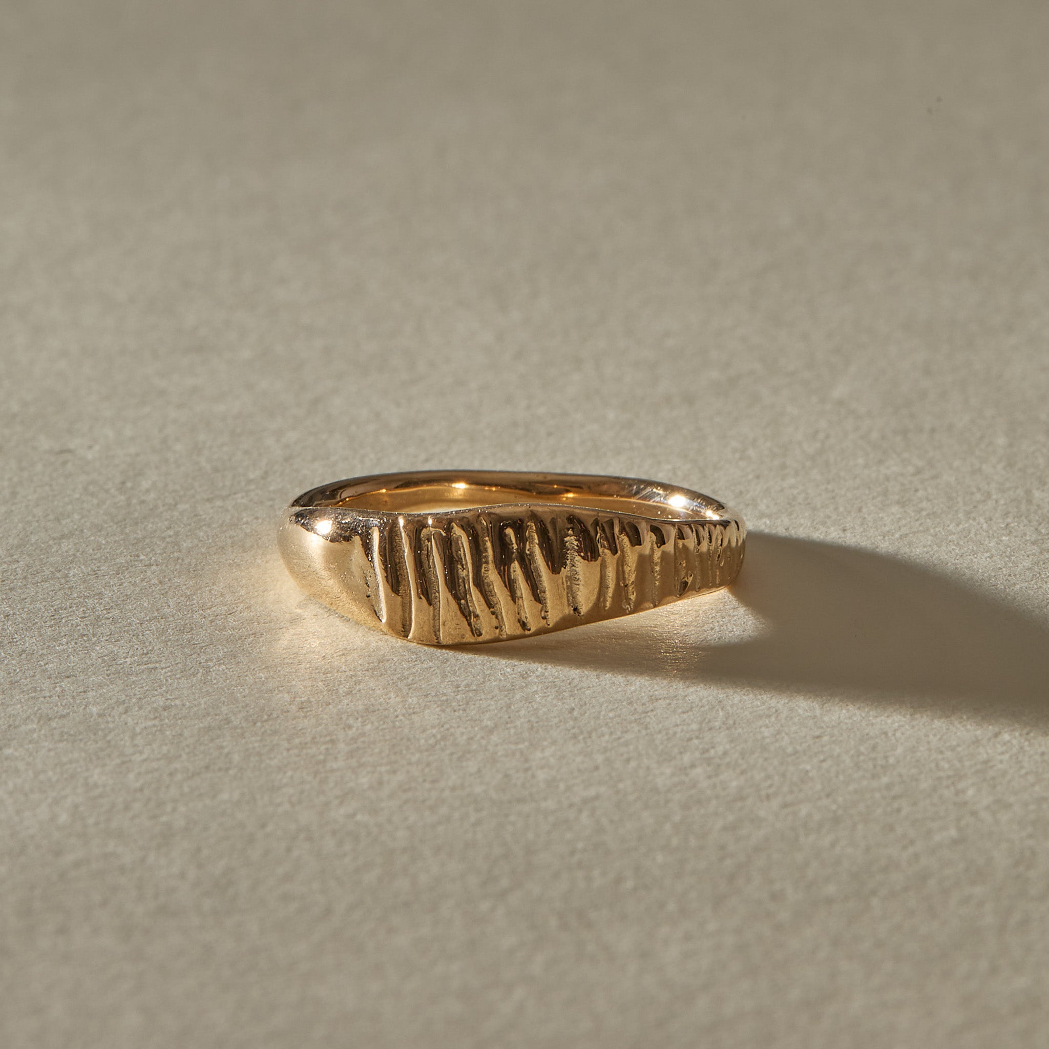 Smoke Ring in solid recycled gold, with flickering vertical lines carved across a sculpted signet face, in warm sunlight.