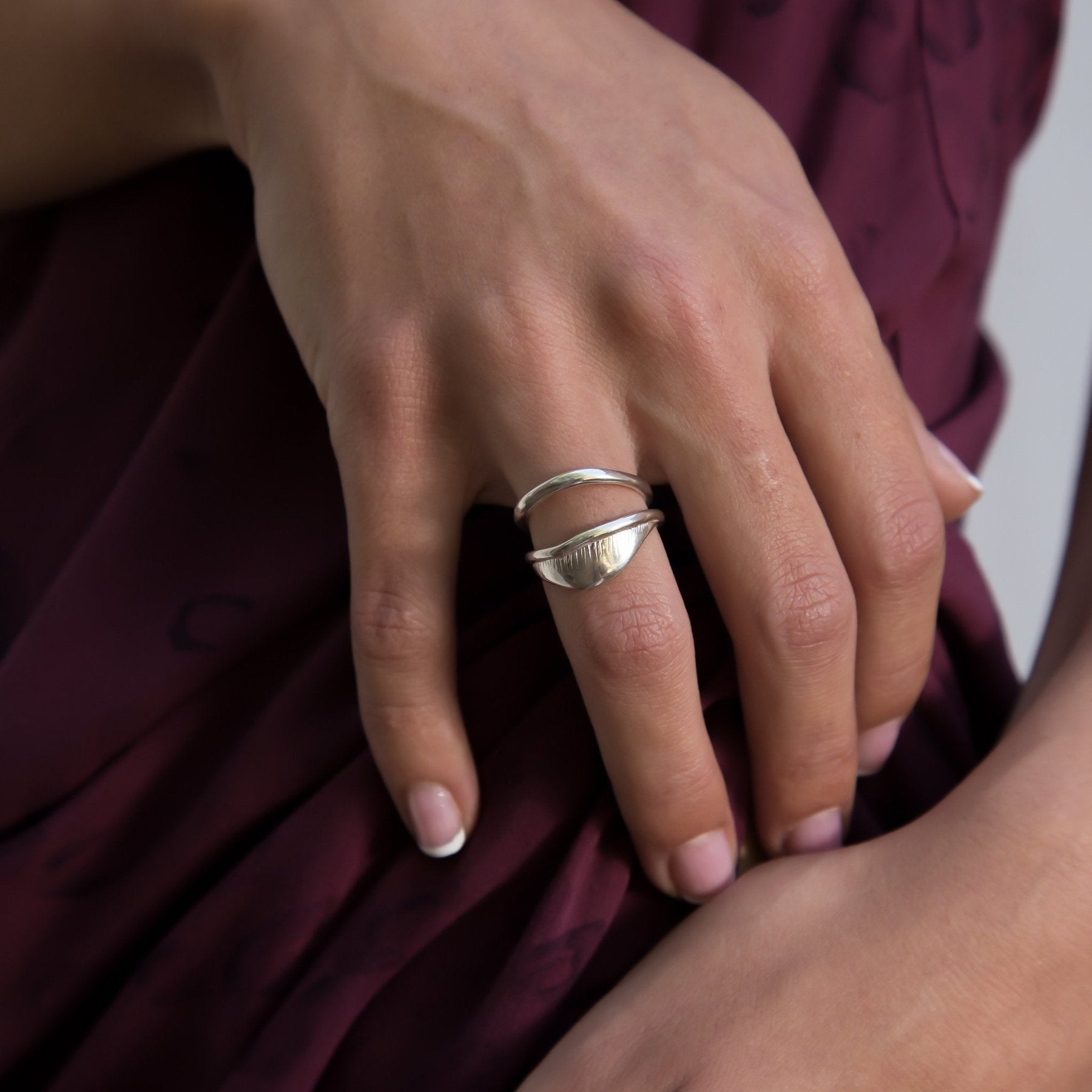 Hand resting on burgundy fabric wearing the Devotions Wave Ring stacked with another ring.