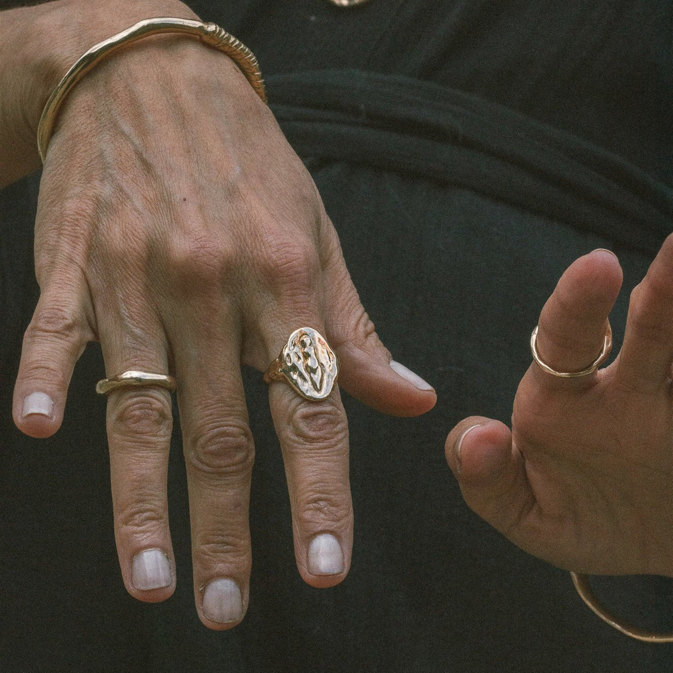 Model's hands against dark fabric wearing the Venus Signet Ring, a carved goddess signet, with organic bangles and rings.