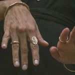 Model's hands against dark fabric wearing the Venus Signet Ring, a carved goddess signet, with organic bangles and rings.